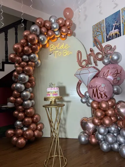 A chic bridal shower decoration featuring a modern white arch backdrop with a "Bride to be" neon sign. The setup is framed by a metallic rose gold and silver balloon arch and includes themed foil balloons like a diamond ring.