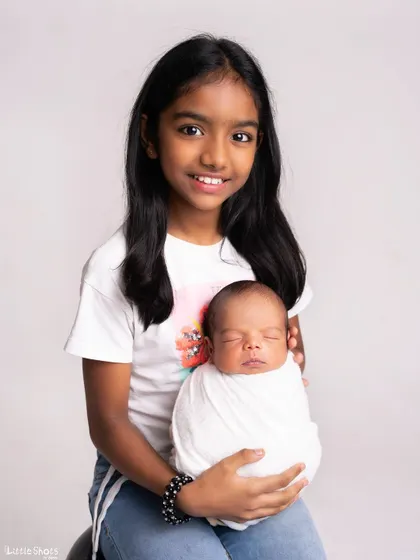 A proud big sister holds her swaddled newborn sibling in this classic studio portrait. Her bright smile shows her excitement at her new role.