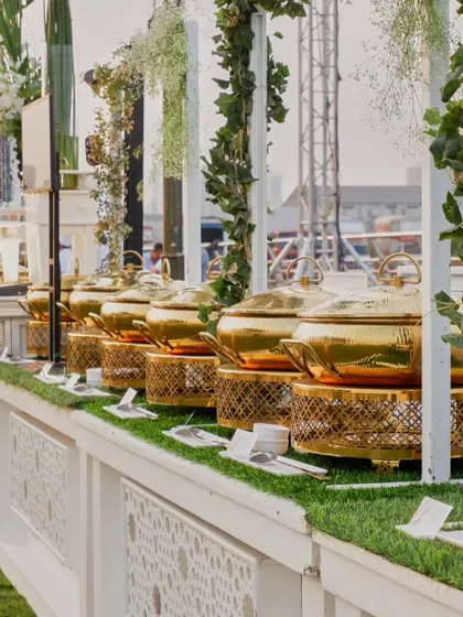 A row of gleaming golden chafing dishes on a pristine white buffet counter. This setup, adorned with fresh greenery, is a picture of elegance and readiness.