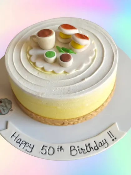 A unique cake designed for a 50th birthday celebration. The top features miniature, handmade fondant models of a cup of tea and Indian sweets, reflecting the client's favorites.