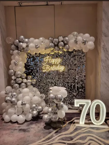 A classic and elegant 70th birthday decoration. The setup features a silver shimmer wall, a white and silver balloon arch, a neon sign, and large marquee numbers.