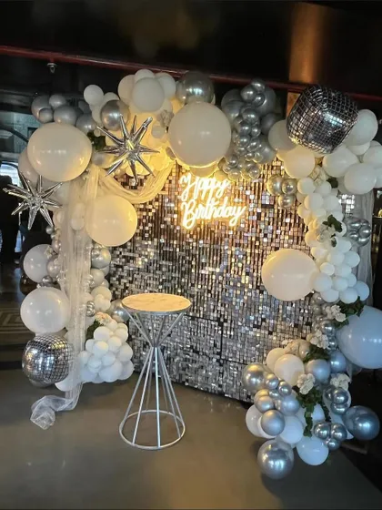 A white and silver disco-themed birthday party. This setup includes a silver shimmer wall, a neon sign, and a beautiful balloon arch with starburst and disco ball accents.