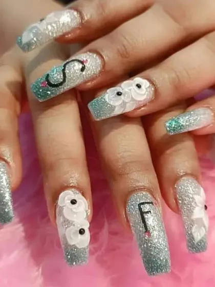 We can personalize your nails with initials or custom designs. This set features long, glittery extensions with 3D flowers and the letters 'S' and 'F'.