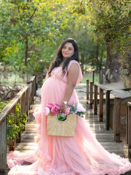 A charming outdoor portrait of a mom-to-be in a pink gown, walking across a wooden bridge while holding a basket of flowers.