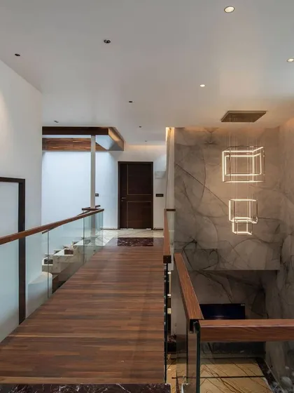 This bridge corridor connects two sections of the upper floor, overlooking a double-height space. The combination of wood flooring, glass railings, and a modern cubic chandelier creates a sophisticated transition area.