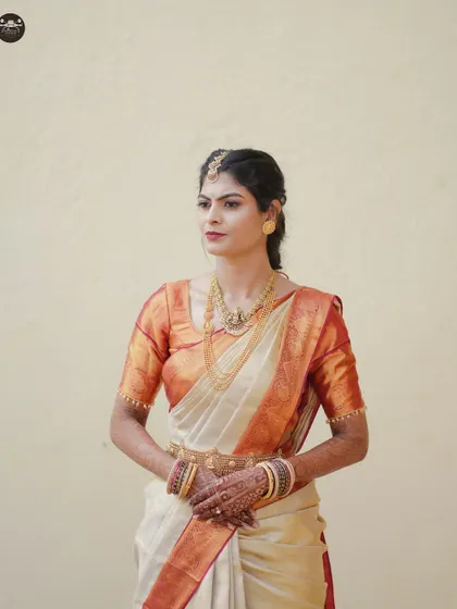 A classic bridal portrait of a South Indian bride in a beautiful off-white and orange silk saree. She is adorned with traditional gold jewelry, including a kamarbandh and maang tikka, showcasing her elegance.