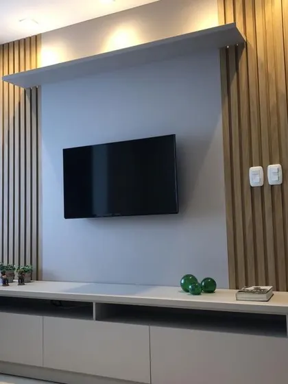 A sleek and modern TV unit design. This unit combines a simple grey console with natural wood-fluted paneling and an overhead shelf with spotlights, creating a stylish focal point for the living room.