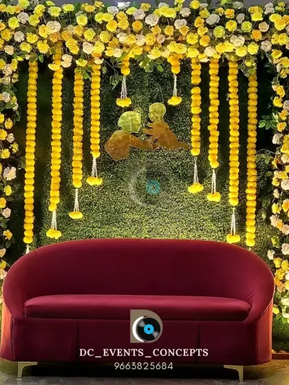 A vibrant baby shower stage with a backdrop of green turf and yellow marigold garlands. The red sofa adds a pop of color, creating a cheerful and festive setting to celebrate motherhood.