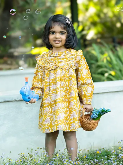 A little girl in a yellow floral dress holds a bubble wand and a basket of flowers. An idyllic outdoor photo, full of innocence and charm.