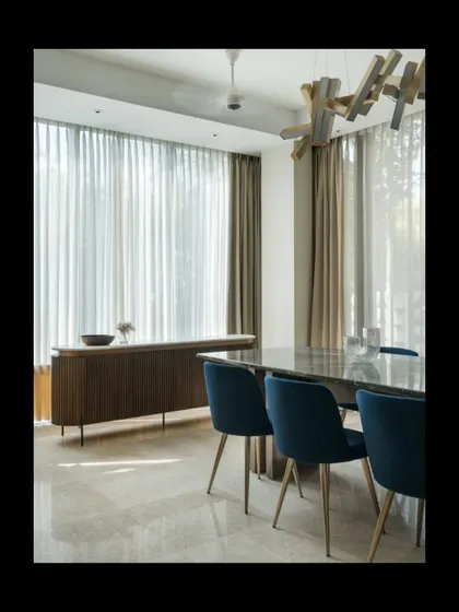 The dining area is designed as a bright, airy space for gathering. A sculptural wooden light fixture hangs above the marble-topped table, while sheer curtains diffuse the sunlight, creating a soft glow. The material palette of wood, stone, and rich blue upholstery adds layers of texture and warmth.