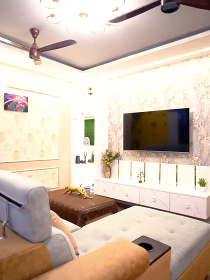 A cozy and luxurious living room featuring a comfortable L-shaped sofa, elegant wallpaper, and a modern TV unit. The false ceiling with dual-tone lighting adds warmth and sophistication to the space.