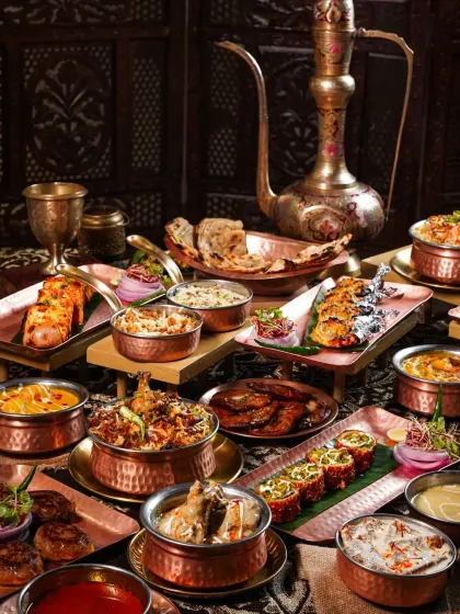 A lavish spread of Awadhi cuisine, featuring biryani, kebabs, and curries in copper and brass vessels. The dark, opulent styling with an antique ewer evokes the royal heritage of Lucknowi food.