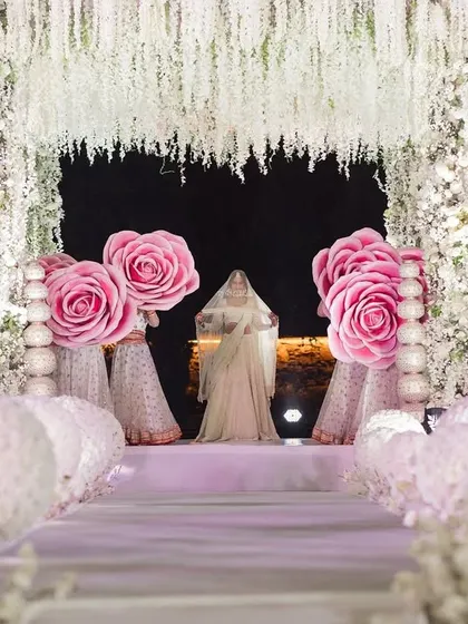 A stunning vision walking down the aisle. The path is lined with white floral spheres and giant pink roses, leading to a mandap draped in hanging white flowers.