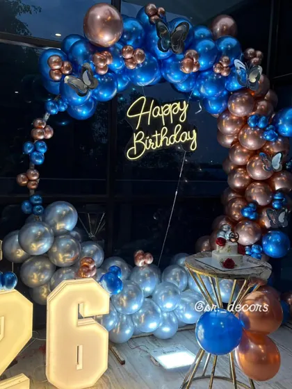 A stunning setup for a 26th birthday, featuring a circular balloon arch in blue, silver, and rose gold, a neon sign, and light-up numbers.