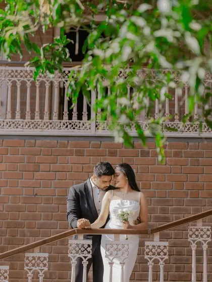 An intimate moment on a balcony, framed by lush green leaves. This photo blends modern architecture with natural elements for a soft, romantic urban portrait.