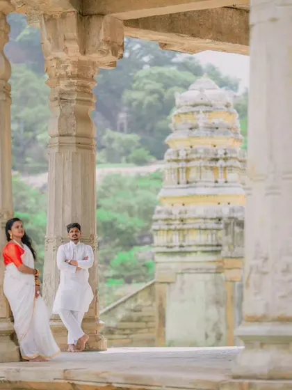 A wide shot of a couple posing amidst the stone pillars of a historic temple, with the gopuram visible in the background. This composition emphasizes the grandeur of the location.