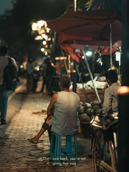 An elderly man sits on a stool on the pavement, a quiet observer of the city's nightlife. His posture tells a story of rest after a long day.