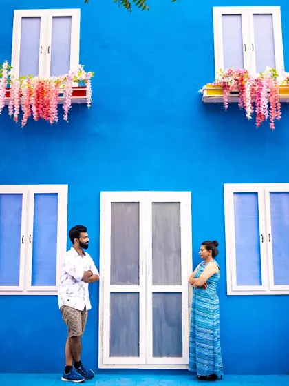 A couple stands against a bright blue wall with white windows, creating a striking and modern pre-wedding photograph. The bold colors make for a visually interesting composition.