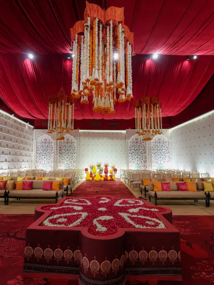 A grand hall set up for a traditional ceremony, with a rich red carpet, a ceiling of red drapes, and chandeliers made of floral garlands.