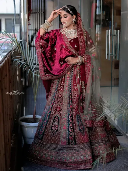A full-length outdoor shot of a bride in a heavily embroidered velvet lehenga. The pose showcases the entire outfit, from the flowing skirt to the rich velvet shawl.