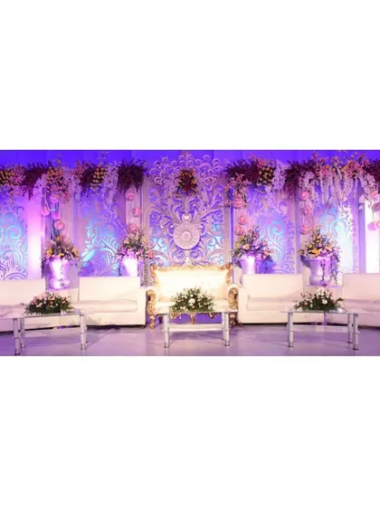 An exceptionally detailed and luxurious reception stage. The intricate white and gold carved backdrop, illuminated by soft purple lighting and adorned with flowers, provides a truly regal setting for the couple.