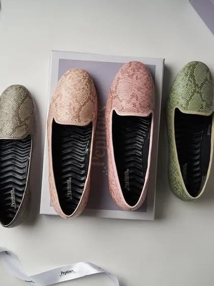 A top-down flat lay for Pepitoes, showcasing their snakeskin-textured loafers. The composition with the box and ribbon gives it a "new shoes" feel, and the soft lighting is perfect for showing off the pastel colors.