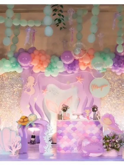 A full stage setup for a mermaid party, featuring a scalloped dessert bar, character cutouts, and a ceiling of hanging balloons that look like bubbles and seaweed.