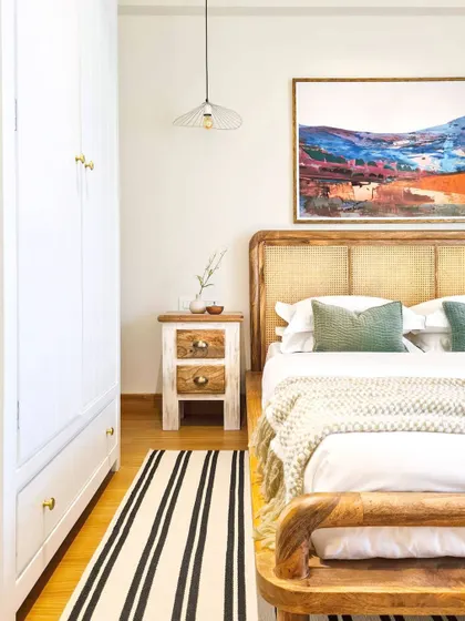 For the guest bedroom in the Breeze Home, I focused on creating a warm and welcoming atmosphere. The bed frame with its curved, caned headboard is the centerpiece, complemented by a colorful piece of art and a simple striped runner rug.