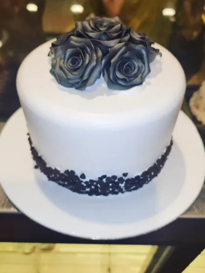 A striking and minimalist cake with a clean white fondant finish. The design is elevated by a cluster of beautifully crafted black sugar roses on top.