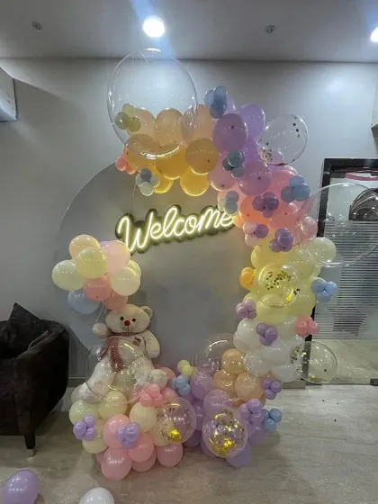 This pastel-themed welcome decoration features a circular backdrop with a neon sign, a cute teddy bear, and an organic balloon garland. It's a perfect, gentle theme for a newborn.