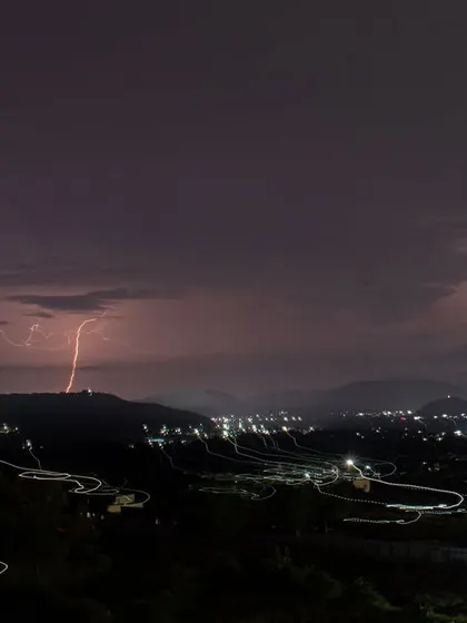 A long-exposure shot captures a lightning strike over Pune, combined with the light trails of vehicles on a winding road.