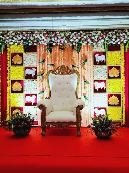 A traditional Seemantham backdrop with a checkerboard pattern of flowers and fabric. The design includes auspicious cow motifs and lotus cutouts against a rich red curtain.