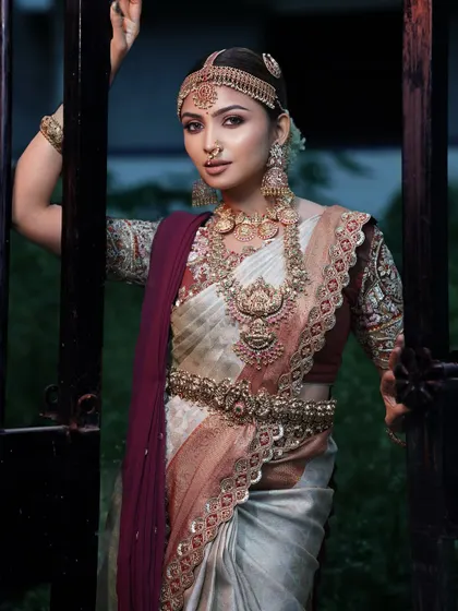 The bride posing at a gate, showcasing the full traditional attire. The makeup is designed to complement the heavy jewellery and intricate saree, creating a cohesive and royal look.