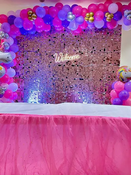 A unicorn-themed welcome party setup with a pink sequin wall and a unicorn foil balloon. The cake table is draped in pink tulle.