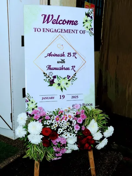 A classic welcome sign for an engagement ceremony, clearly stating the couple's names and event date. The simple floral arrangement at the base adds a touch of freshness and celebration.