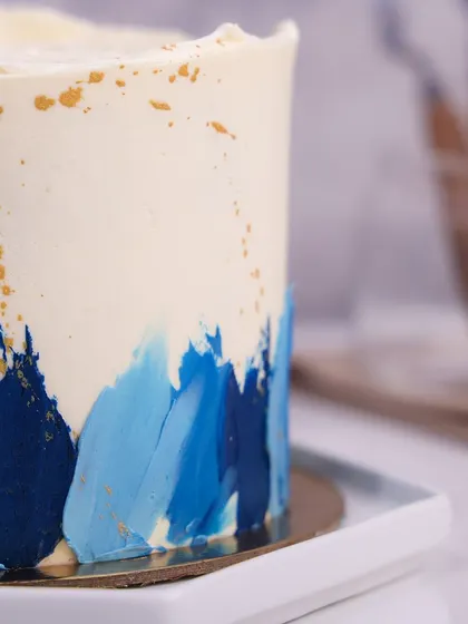We create a piece of modern art with this abstract blue buttercream cake. Strokes of various blue shades are painted onto a white canvas, accented with gold splatters and topped with piped rosettes, making it a stylish and happy choice for a Father's Day celebration.
