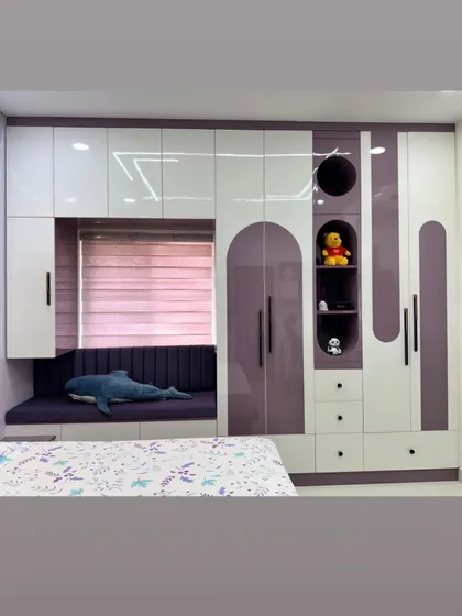 For this kids' room, we designed a multi functional wardrobe unit in purple and white. It includes a cozy seating nook by the window, open shelves for toys, and plenty of cabinet space.