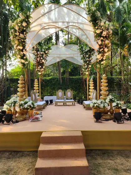 This unique mandap design for an outdoor wedding features a modern arched roof structure draped in sheer fabric and adorned with clusters of white flowers, creating an airy and elegant space.