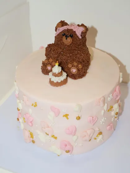 An adorable teddy bear themed birthday cake. This one features a cute, fuzzy buttercream bear wearing a pink bow, sitting next to its own tiny birthday cake.