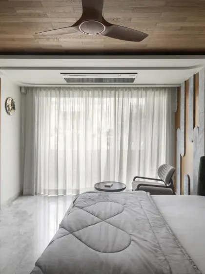 This view of the father's bedroom in the Slender House shows the warm wooden ceiling and full-height sheer curtains. The design prioritizes comfort and relaxation, with a connection to the community via the front balcony.