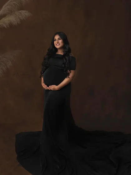 A full-length portrait showing the dramatic sweep of the black gown. The simple, textured backdrop and pampas grass add a touch of warmth and modernity to this classic studio shot.