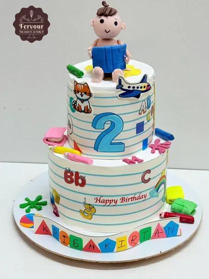 A school-themed cake for a 2nd birthday. The tiers are designed to look like notebook paper, decorated with letters, numbers, and a cute fondant baby reading a book on top.