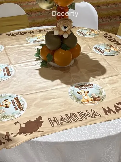 A close-up of a guest table at the Lion King party, featuring a custom 'Hakuna Matata' tablecloth and themed placemats. The centerpiece is a fun balloon arrangement topped with a small lion toy.