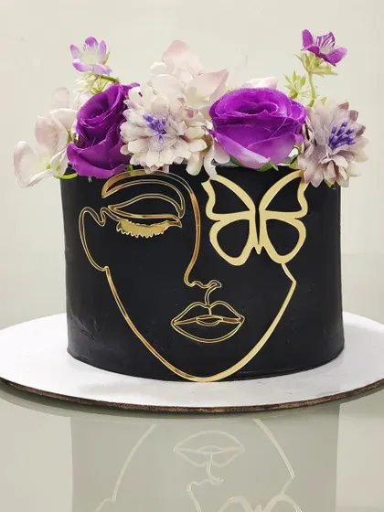 A striking black cake with a gold abstract face and butterfly design. Topped with a crown of fresh purple flowers, this cake is a perfect example of bold, artistic, and modern cake design.