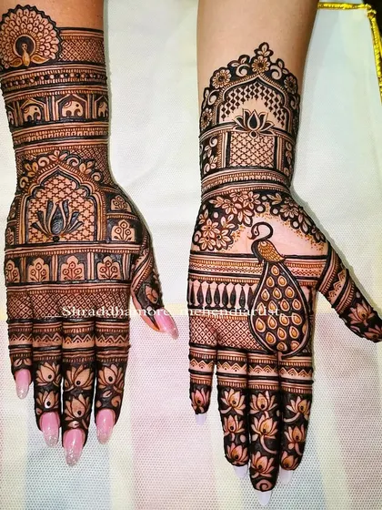 This design for Shivani features a beautiful mix of architectural elements and nature. One hand has a majestic peacock, while the other showcases a lotus inside a jharokha, with elephant motifs on the wrist.
