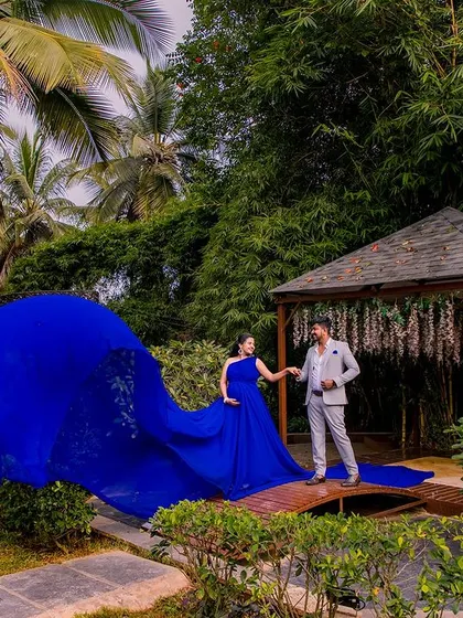This wide shot showcases the incredible length of the flowing blue maternity gown. Set against a wooden bridge and lush bamboo, the fabric creates a stunning visual that fills the frame with color and movement.