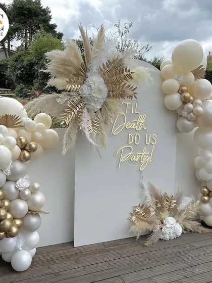 A fun and chic outdoor party setup with a "Til Death Do Us Party" neon sign. The white and gold balloon decor with pampas grass is perfect for a bachelorette or milestone birthday.