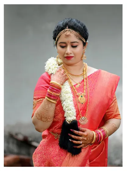 A classic three-quarter portrait of a bride in a red saree, holding a garland of fresh jasmine flowers.