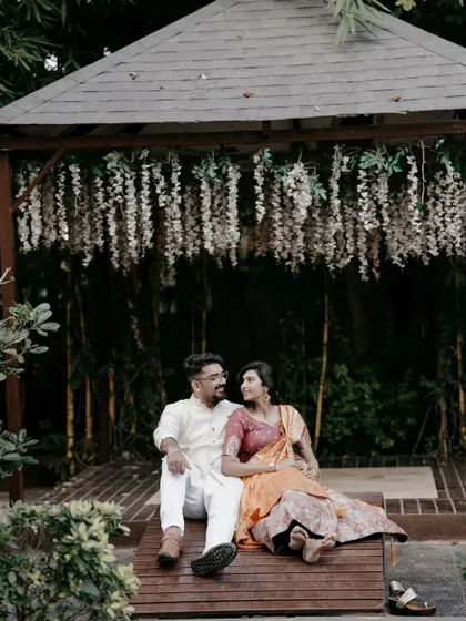 A romantic shot of a couple in traditional wear, relaxing under a gazebo adorned with hanging white flowers.