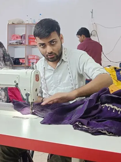My team works diligently to bring each design to life. Here, a tailor is focused on stitching a purple garment, ensuring every seam is perfect.